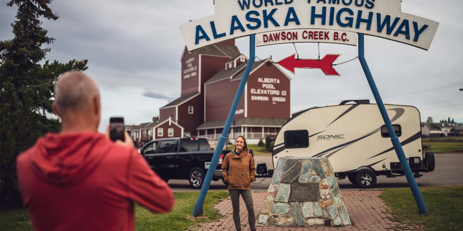 couple taking photo by RV at alaska highway sign