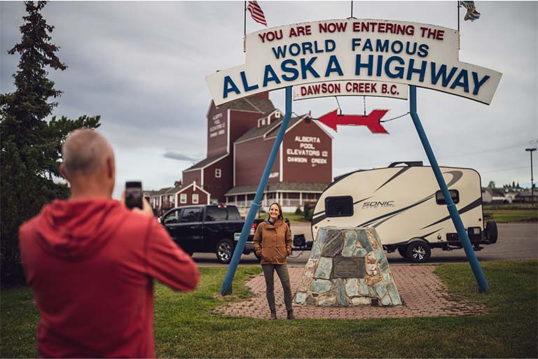 couple taking photo by RV at alaska highway sign