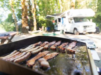 Griddle with bacon cooking at a campsite with a Class C motorhome in the background