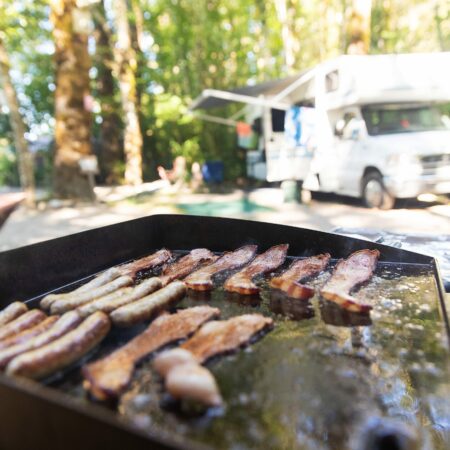 Griddle with bacon cooking at a campsite with a Class C motorhome in the background