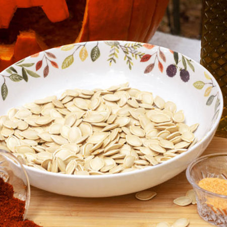 A bowl of pumpkin seeds placed on a table with seasonings, tobasco sauce, and a jackolantern